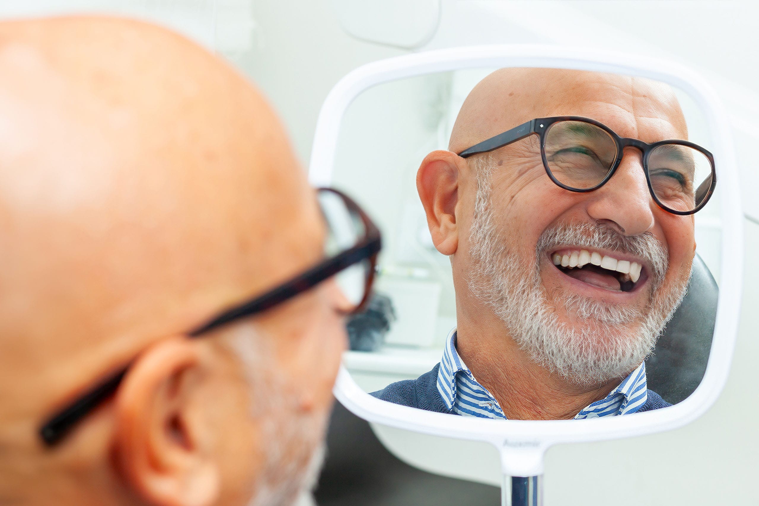 Older man smiling while checking his teeth in a handheld mirror during a dental visit